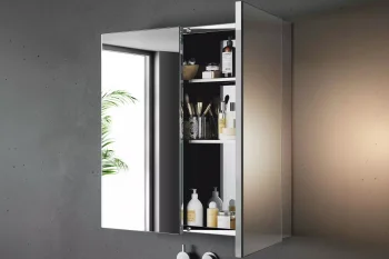 Chrome bathroom cabinet with mirrored doors, shelves of toiletries inside; grey textured wall, tap below, and a reflected window with a plant.