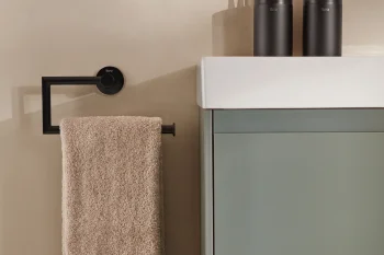 Beige bathroom wall with a black square towel rail holding a beige towel; white sink top above a sage cabinet, with dark cylindrical canisters and toothbrushes on the counter.