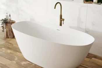 White freestanding bathtub on wooden floor with brass faucet, marble wall tiles and lush greenery visible in the background.