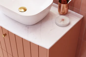 White vessel sink with a gold drain sits on a marble countertop atop a pink paneled vanity; a gold toothbrush holder and round soap sit by pink tiles.