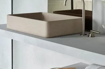 Beige rectangular vessel sink on a grey countertop with a curved chrome faucet; mirrors behind and a shelf below with white towels and a small dish.