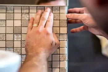 Close-up of hands placing small textured square tiles on a grid panel, with a ring on one finger and another person guiding the board.