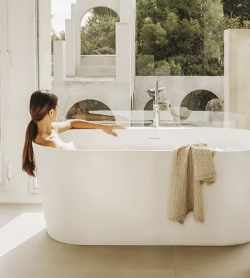 Person sits in a white freestanding bathtub in a bright room with large windows; a beige knitted towel drapes the tub edge, wooden stool nearby, arches outside.