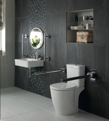 Modern bathroom with glass shower, grey tiles, wall-mounted sink and circular mirror, toilet with grab bars, open shelves, vase of flowers by window.
