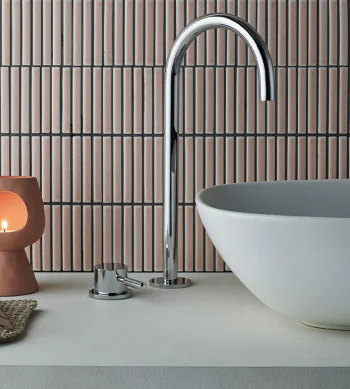 Modern bathroom with oval white basin and chrome curved faucet, terracotta candle holder with a lit candle, woven mat and seashell on countertop.