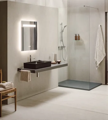 Minimalist bathroom with concrete walls, a backlit mirror, black sink on a floating counter, glass shower and a wooden chair with folded towels.