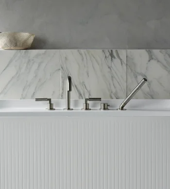 White ribbed freestanding bathtub with chrome fixtures in a marble-tiled bathroom; grey wall above and a woven basket on a ledge.