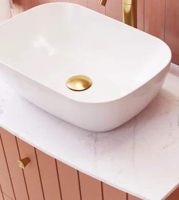 White vessel sink on a pale marble countertop with pink-tiled walls; gold faucet and drain, copper toothbrush holder, and a round marble soap dish.