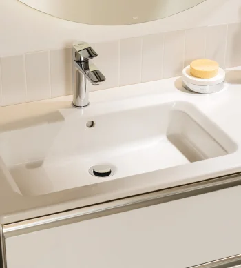 White bathroom sink with chrome faucet, soap on a dish, and a toothbrush holder with two wooden toothbrushes on a glossy white counter.