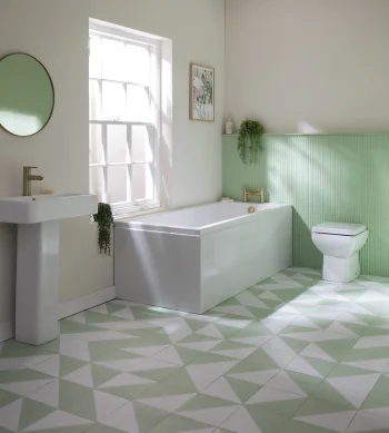 Bright bathroom with a white pedestal sink, freestanding bathtub and toilet, pale green ribbed wall panels, mint and white geometric tiled floor, round mirror, window and plants.