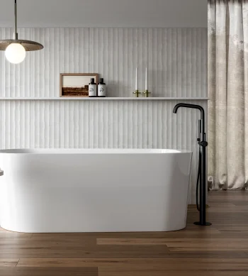 Minimalist bathroom with a white freestanding tub, black floor tap, wood floors, textured white wall, table with dried stems, and beige curtains.