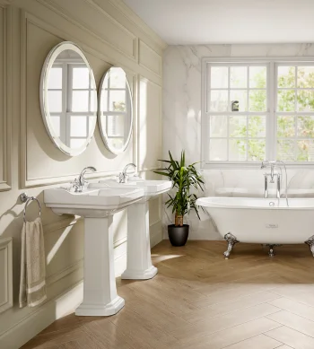 Bright bathroom with a freestanding clawfoot tub by a large window, two pedestal sinks with mirrors, marble walls, a plant and a herringbone floor.