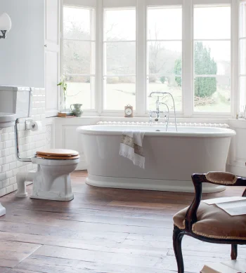 Bright vintage bathroom with a freestanding white bath by a bay window, pedestal sink, toilet with a wooden seat, and ornate dark wood armchair.