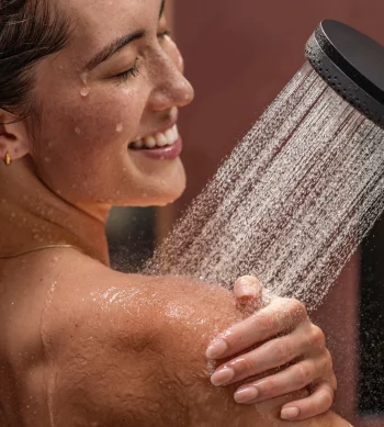 Close-up of a person with wet hair smiling under a handheld shower, water cascading over their shoulder as they wash.