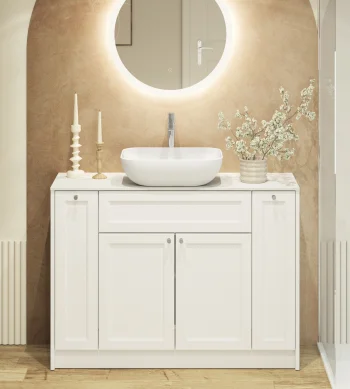 White bathroom vanity with round backlit mirror, vessel sink, and a vase of white blossoms on a beige wall; candles left, glass shower right.