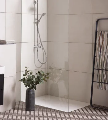 Modern bathroom with a glass shower enclosure, chrome rainfall and handheld shower, beige tiles, a patterned towel rack, and a vase of eucalyptus.