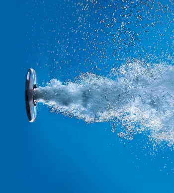 Water spurts from a circular nozzle on the left, forming a frothy, bubbly plume across a blue background.