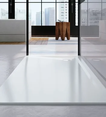 Modern interior with glass walls and city skyline view; white rectangular shower tray in the foreground, grey seating to the left, and a wooden stool.