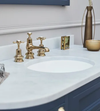 Gold brass faucet with two handles on a white marble bathroom sink, mounted in a blue vanity; a tall bronze vase and small bowl sit to the right.