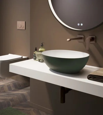 Modern bathroom with a round backlit mirror, white floating counter, green bowl sink, wall-mounted tap, soap bottles, towel, and herringbone floor.