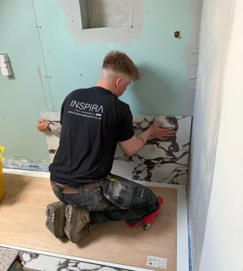 Person kneels on plywood floor, placing a marble-pattern tile along a pale green bathroom wall during renovation; wearing a black Inspira shirt.