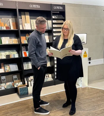 Two people in a tile showroom study a CA' PIETRA brochure; tall black display shelves behind them hold various tile samples.