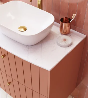 White vessel sink with a gold drain sits on a marble countertop atop a pink paneled vanity; a gold toothbrush holder and round soap sit by pink tiles.