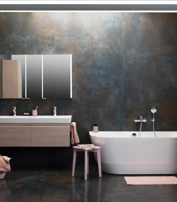 Modern bathroom with dark, metallic walls and a wall-mounted wooden double vanity with two sinks and a backlit mirrored cabinet; a white freestanding tub, pink towels and stool.