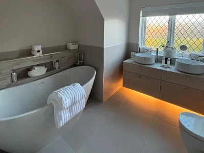 Modern bathroom with a freestanding tub left and a double vanity with round vessel sinks right, warm under-cabinet lighting and a lattice window.