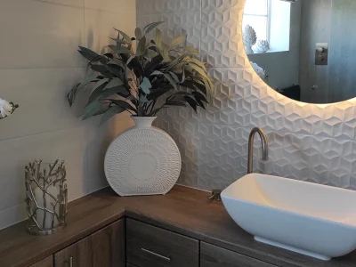 Modern bathroom vanity with a white vessel sink, curved faucet, round backlit mirror, 3D geometric tile wall, and potted plants on a wooden countertop.