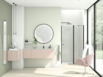 Bright, minimalist bathroom with pale green walls, a pink floating vanity, round mirror, a vase of flowers, glass shower, and a pink lounge chair by a large window.