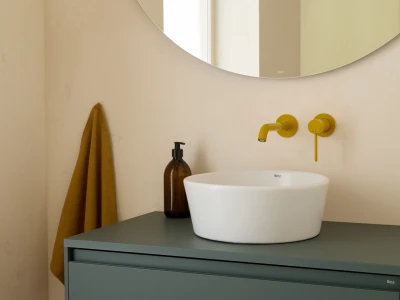 Round mirror above a pale wall; a white vessel sink sits on a blue-green cabinet with a yellow wall-mounted tap and lever, a brown soap dispenser, and a mustard towel.