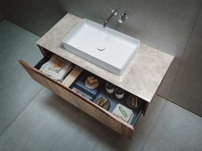 Top‑down view of a modern bathroom vanity with a white rectangular vessel sink on a marble countertop; an open drawer reveals towels and toiletries.