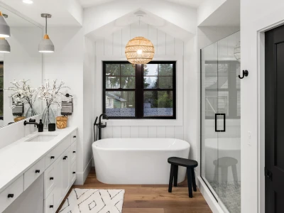 Bright white bathroom with a freestanding tub by a black-framed window, glass shower, long white vanity, woven pendant light, and a black stool on wood floor.