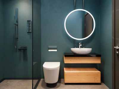 Modern bathroom with teal walls, a round backlit mirror, white vessel sink on a black countertop above timber drawers, wall-hung toilet and glass shower.