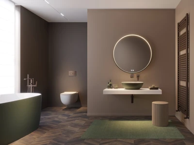 Contemporary bathroom with dark taupe walls, a green freestanding tub, wall-hung toilet, circular backlit mirror, white floating counter with an olive-green basin, ribbed stool, and chevron wood floor.