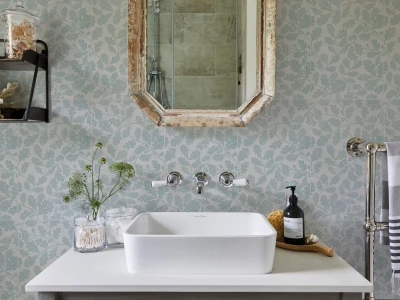 Light-blue leafy wallpaper, octagonal wooden-framed mirror above a white vessel sink; shelf with jars and coral, soap bottle beside a towel rail.