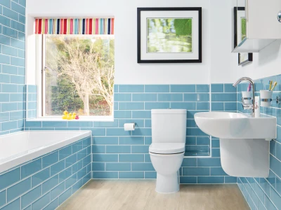 Bright bathroom with blue subway tiles, white toilet and sink, large window with striped blind, rubber ducks on the sill, and a framed leaf photo.