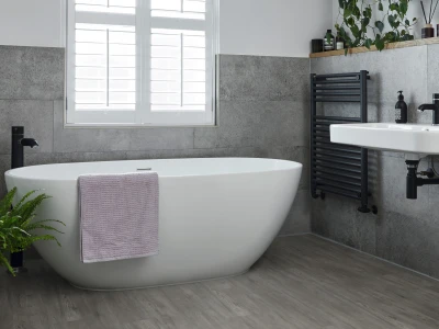 Modern bathroom with a white freestanding tub, lavender towel, black fixtures, grey tiled walls, a wooden shelf, a potted fern and a window with shutters.