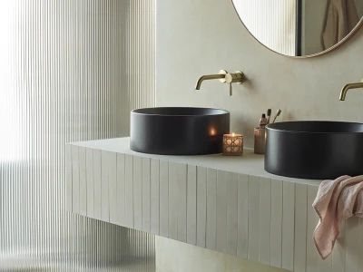 Minimalist bathroom with two matte black round sinks on a light beige countertop, gold wall taps, a round mirror, a lit candle, and a pink towel.