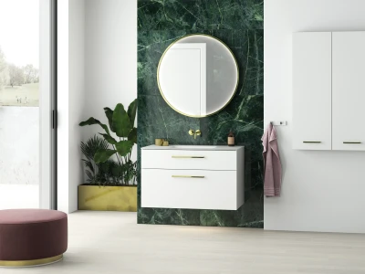 Modern bathroom with white vanity against dark green marble, round gold-framed mirror, white cabinet, gold handles, plants, pink towel, and a window.