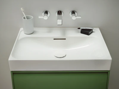 White rectangular bathroom sink with chrome faucet and two handles; toothbrush in a mug on the left, toothpaste and soap on the right, with a green cabinet beneath.