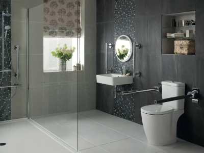 Modern bathroom with glass shower, grey tiles, wall-mounted sink and circular mirror, toilet with grab bars, open shelves, vase of flowers by window.