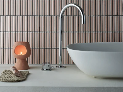 Modern bathroom with oval white basin and chrome curved faucet, terracotta candle holder with a lit candle, woven mat and seashell on countertop.