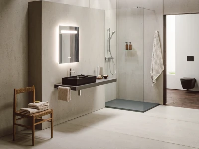 Minimalist bathroom with concrete walls, a backlit mirror, black sink on a floating counter, glass shower and a wooden chair with folded towels.