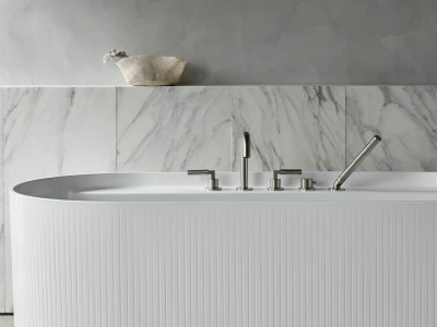 White ribbed freestanding bathtub with chrome fixtures in a marble-tiled bathroom; grey wall above and a woven basket on a ledge.