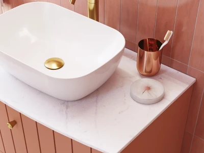 White vessel sink on a pale marble countertop with pink-tiled walls; gold faucet and drain, copper toothbrush holder, and a round marble soap dish.