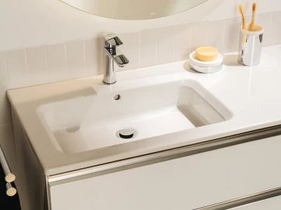 White bathroom sink with chrome faucet, soap on a dish, and a toothbrush holder with two wooden toothbrushes on a glossy white counter.