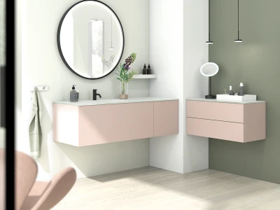 Modern bathroom with pale pink floating vanities, round mirror, black faucet, towel on a hook, vase of flowers, and a freestanding makeup mirror.