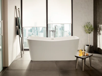 Minimalist bathroom with a white freestanding bathtub, chrome tap, and a city skyline view through large windows. A towel ladder and a stool hold bath oils, soap and a plant.